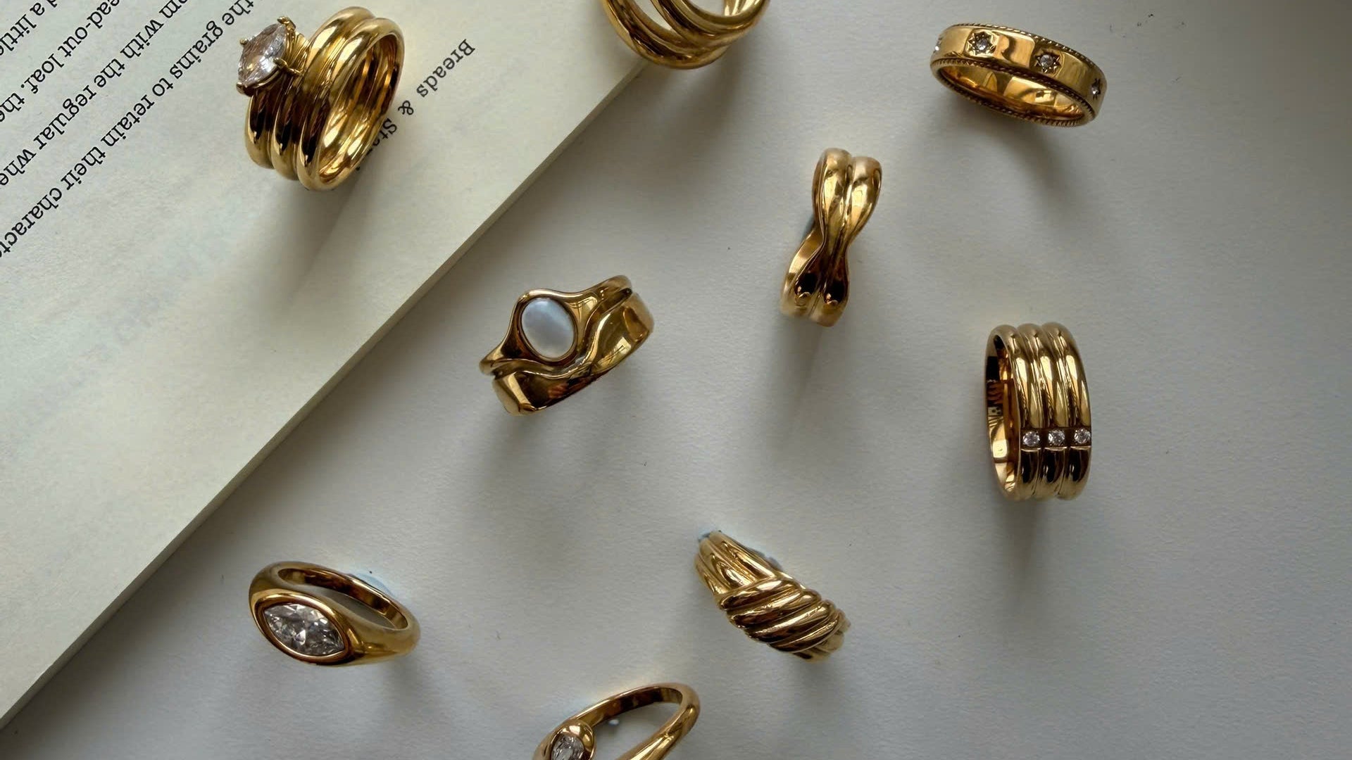 A styled flat-lay of nine gold rings with various twisted, banded, and textured designs, arranged on a light gray surface next to an open book.