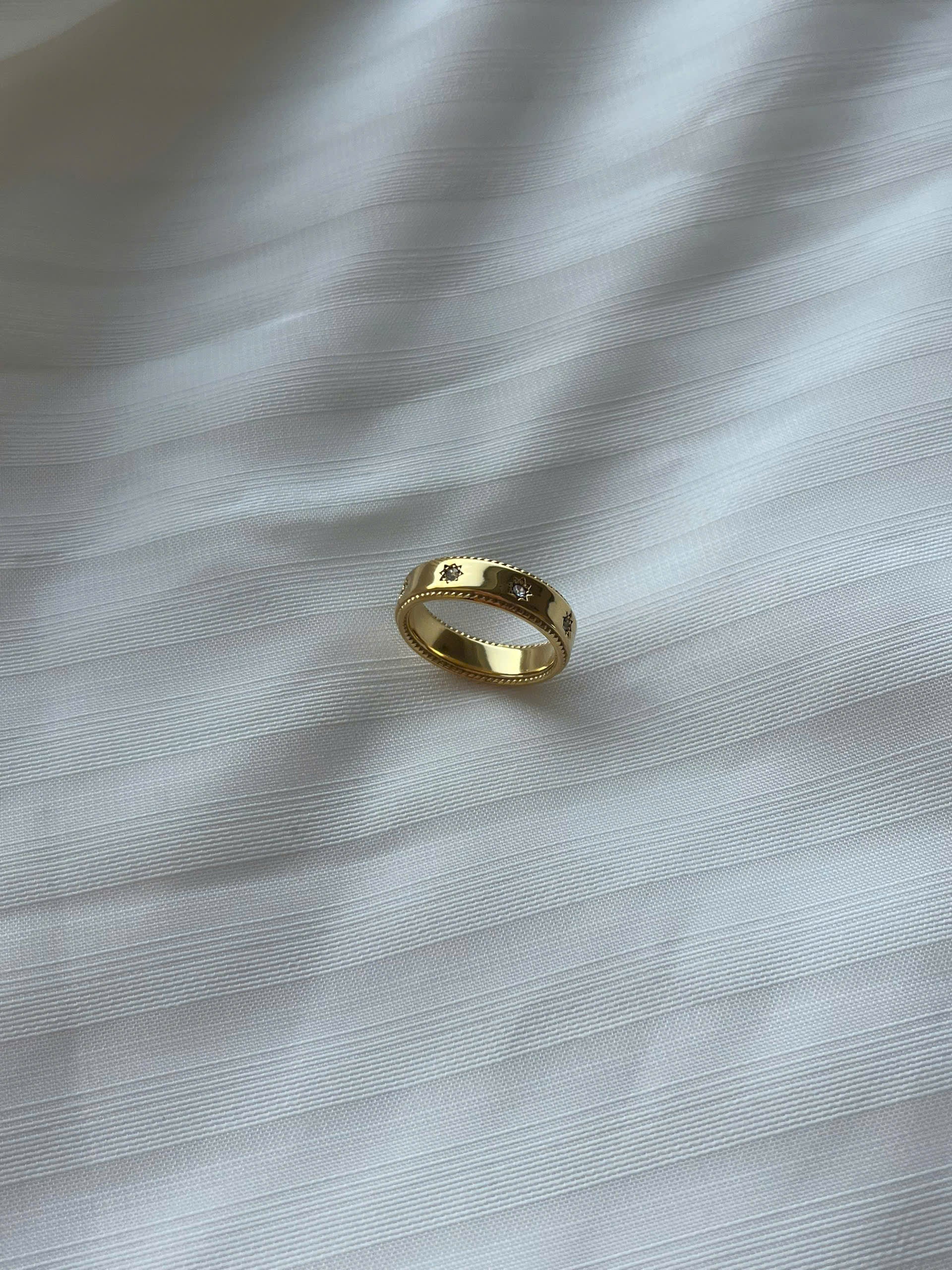 A gold band ring with a textured, beaded outer edge and a smooth inner band, resting on a white textured fabric surface.