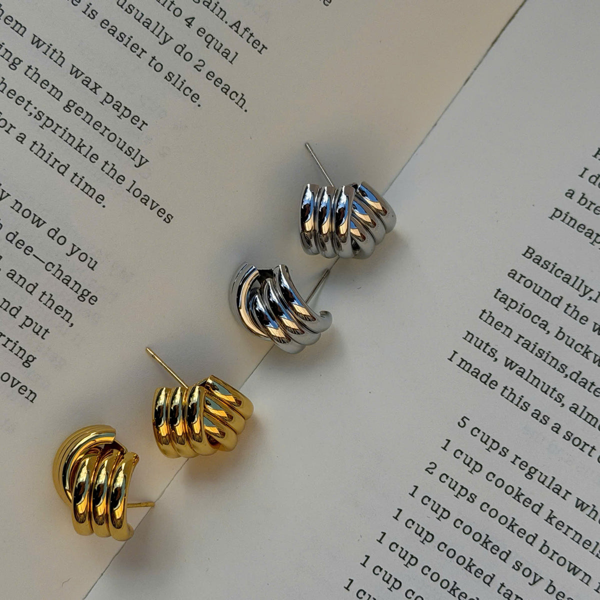 Three small spiral-shaped hoop earrings in silver and gold finishes are arranged on an open book with printed text.