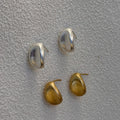 Four small oval stud earrings in polished silver and gold-tone metals, arranged on a textured light gray surface.