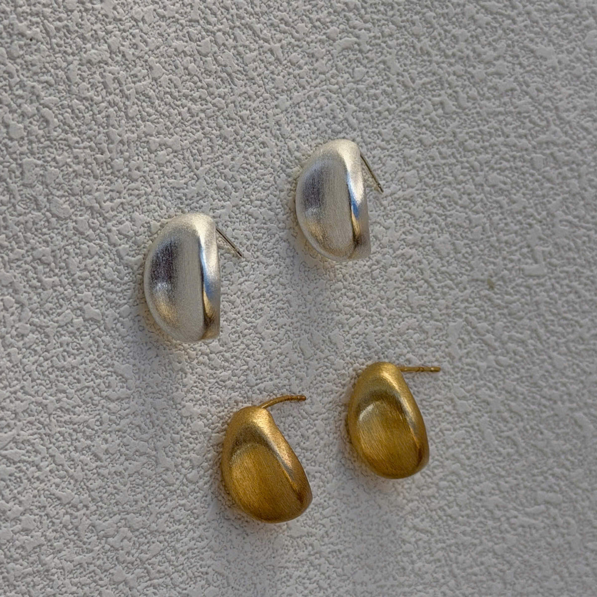 Four small oval stud earrings in polished silver and gold-tone metals, arranged on a textured light gray surface.