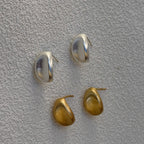 Four small oval stud earrings in polished silver and gold-tone metals, arranged on a textured light gray surface.