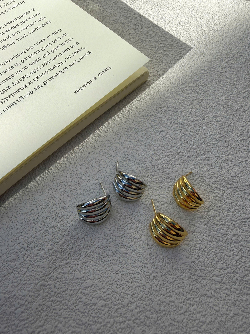 Three small spiral hoop earrings in silver and gold tones arranged on a textured light gray surface next to an open book.