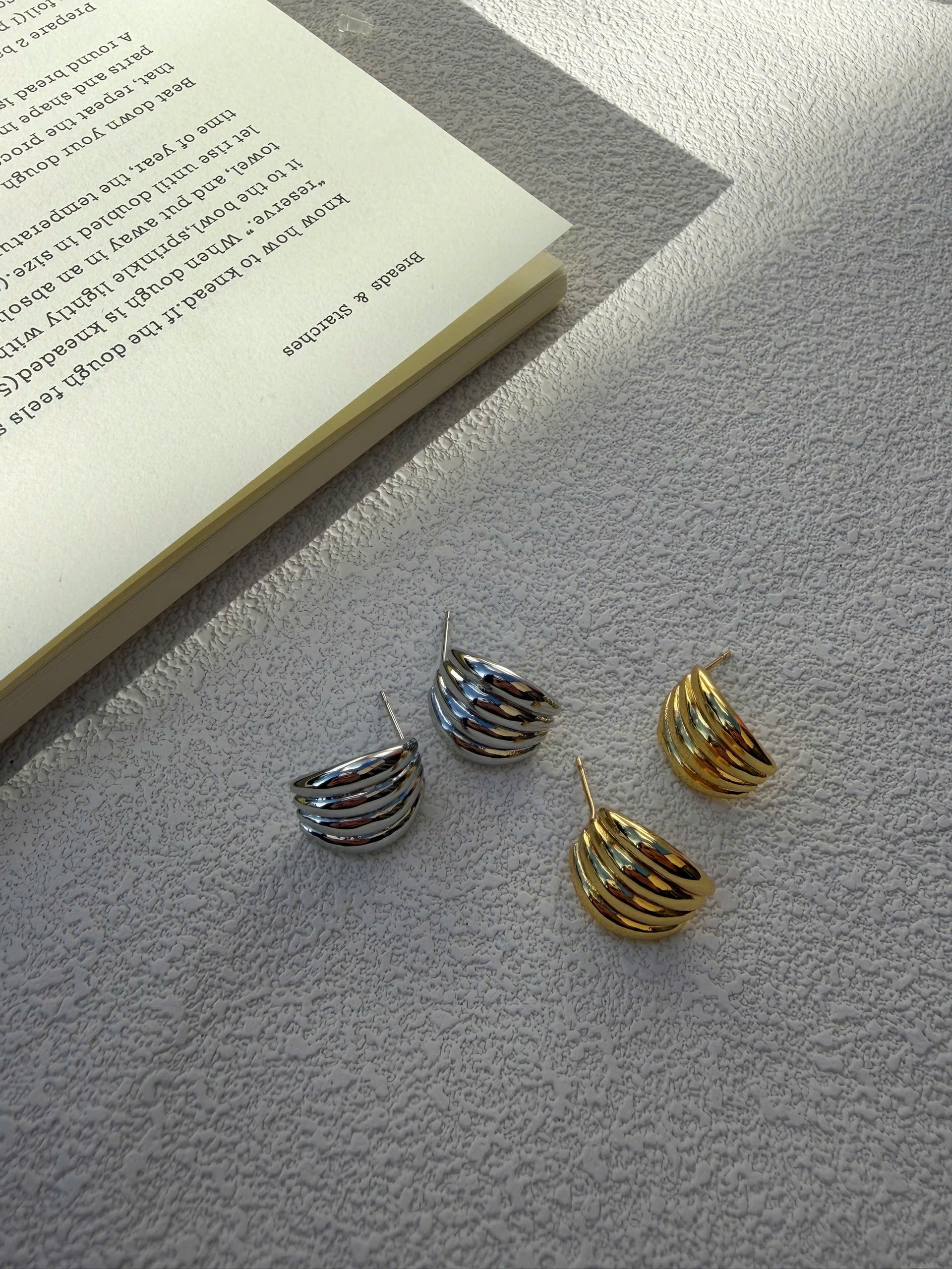 Three small spiral hoop earrings in silver and gold tones arranged on a textured light gray surface next to an open book.