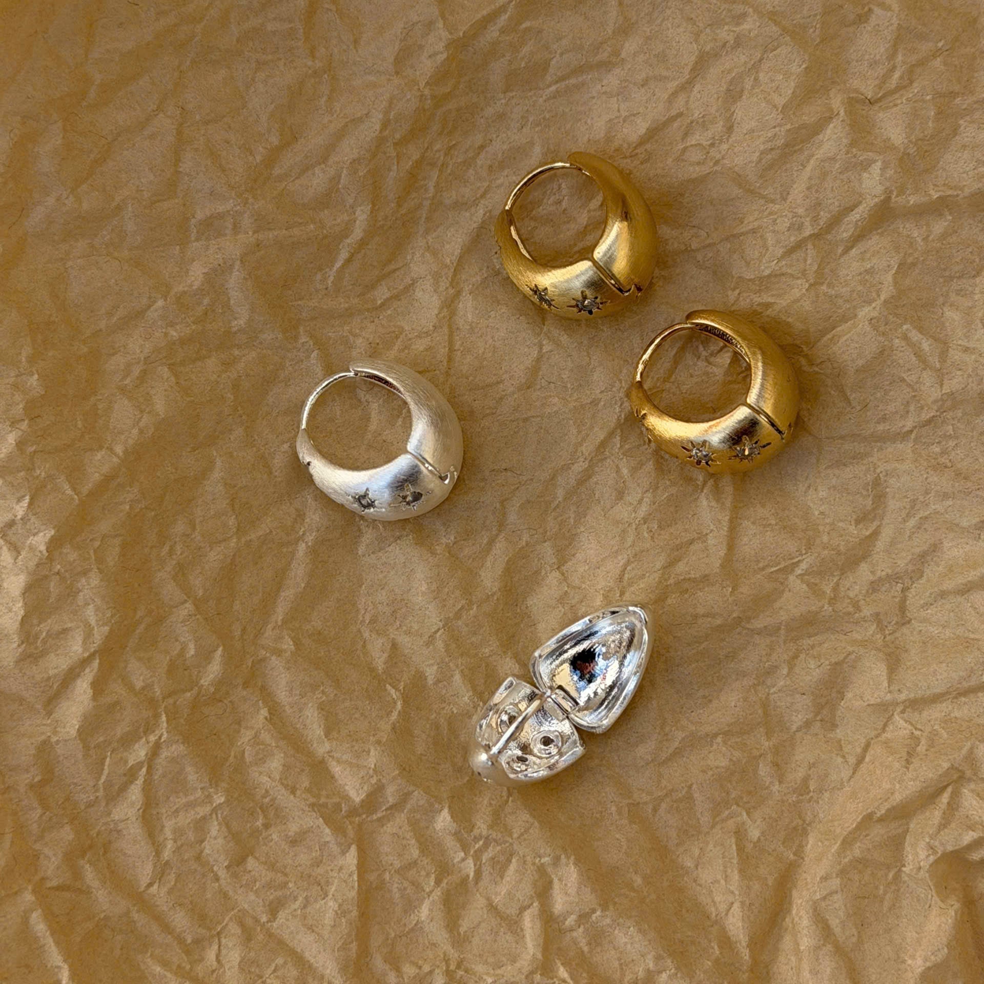Four small metallic jewelry pieces arranged on crumpled brown paper: two gold-toned pieces and two silver-toned pieces, photographed from above.
