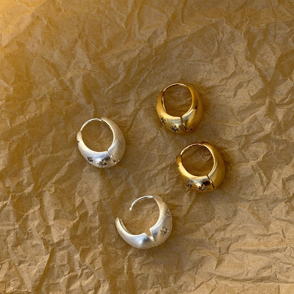Four small metallic hoop earrings in silver and gold finishes arranged on crumpled brown paper.