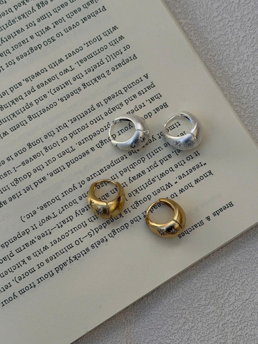Three small hoop-style earrings in silver and gold tones arranged on an open book page.