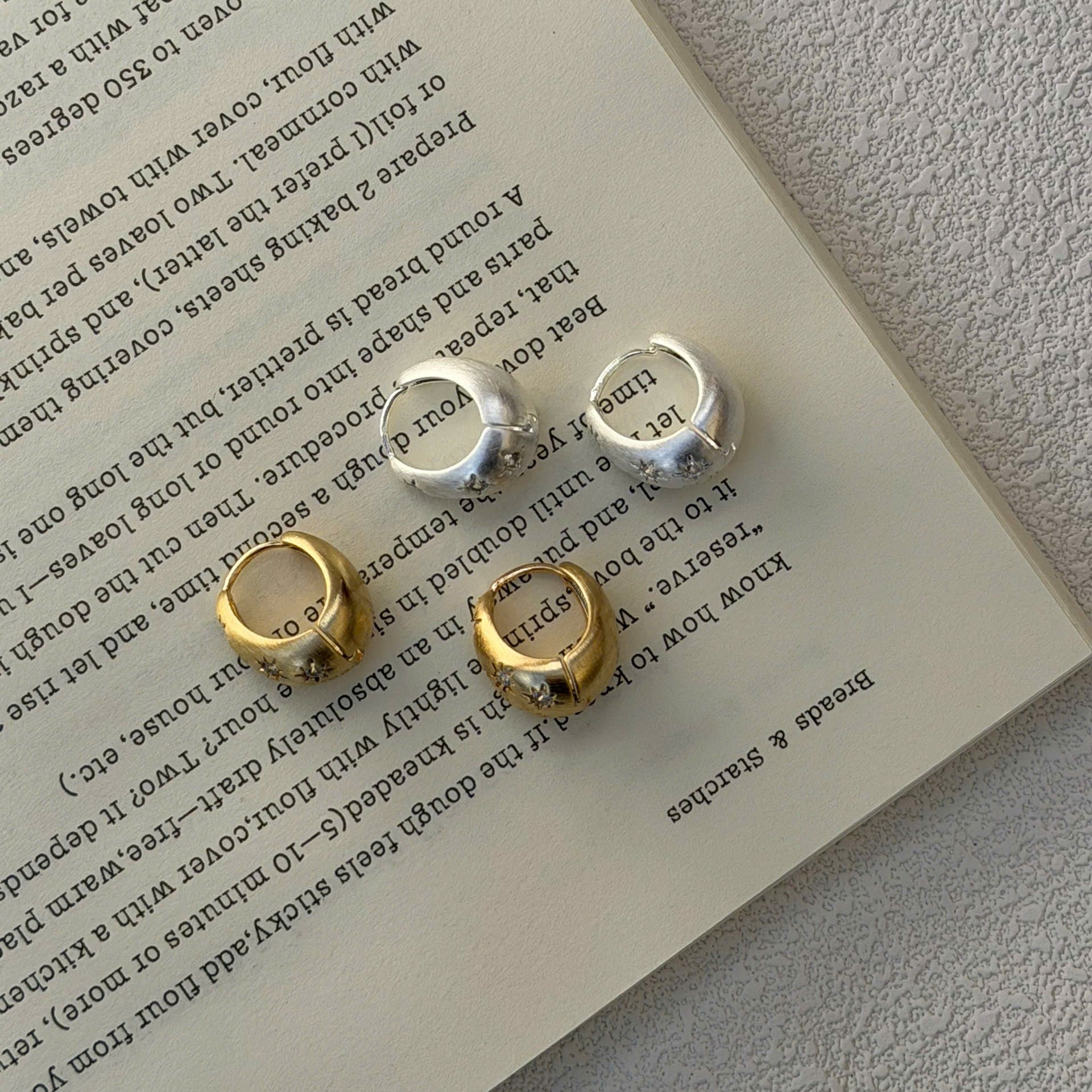 Four small hoop earrings in gold and silver tones arranged on an open book page, with a textured light gray surface visible around the book.