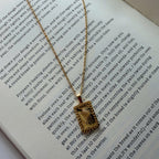 A gold-toned rectangular pendant necklace with a sunburst design hangs over an open book page with printed text.