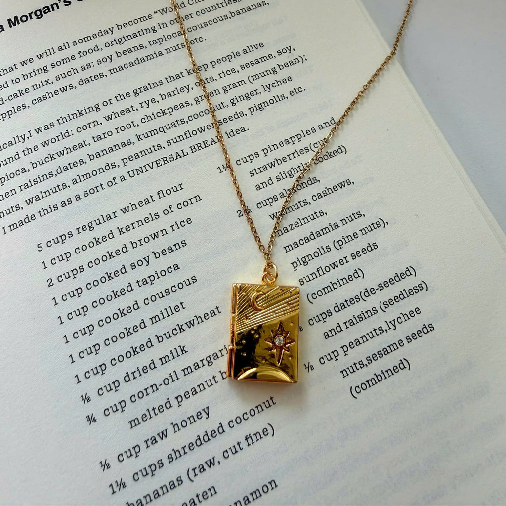 A gold-toned necklace with a rectangular pendant featuring a sunburst design and the text 'Morgan's Global Bread Cake' printed on it, displayed on an open book page.