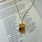 A gold-toned necklace with a rectangular pendant featuring a sunburst design and the text 'Morgan's Global Bread Cake' printed on it, displayed on an open book page.