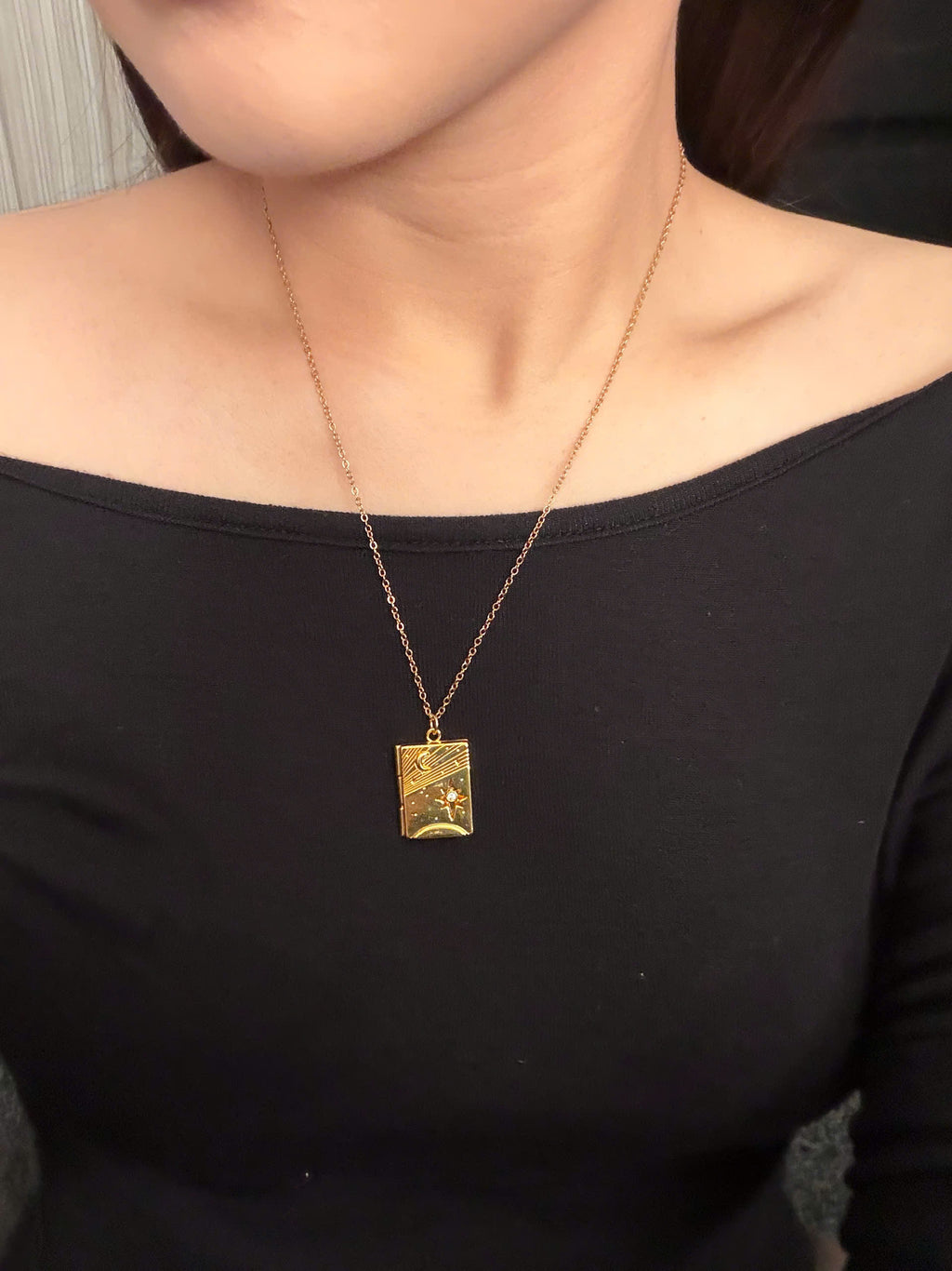 A gold pendant necklace with a textured rectangular charm worn by a model in a black off-the-shoulder top.