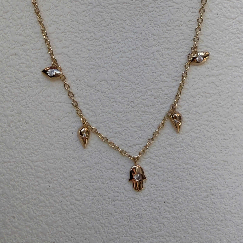 A delicate gold chain necklace featuring small dangling charms, including a central hamsa hand pendant and smaller bird-like accents, displayed on a textured off-white background.