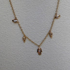 A delicate gold chain necklace featuring small dangling charms, including a central hamsa hand pendant and smaller bird-like accents, displayed on a textured off-white background.