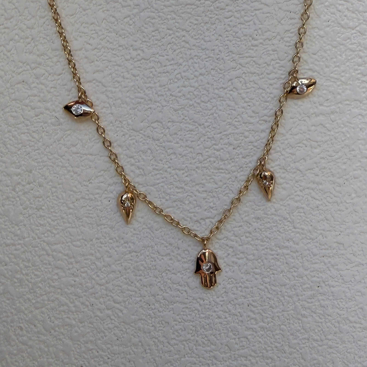 A delicate gold chain necklace featuring small dangling charms, including a central hamsa hand pendant and smaller bird-like accents, displayed on a textured off-white background.
