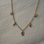 A delicate gold chain necklace featuring small dangling charms, including a central hamsa hand pendant with a blue stone, laid flat on a textured light gray fabric background.