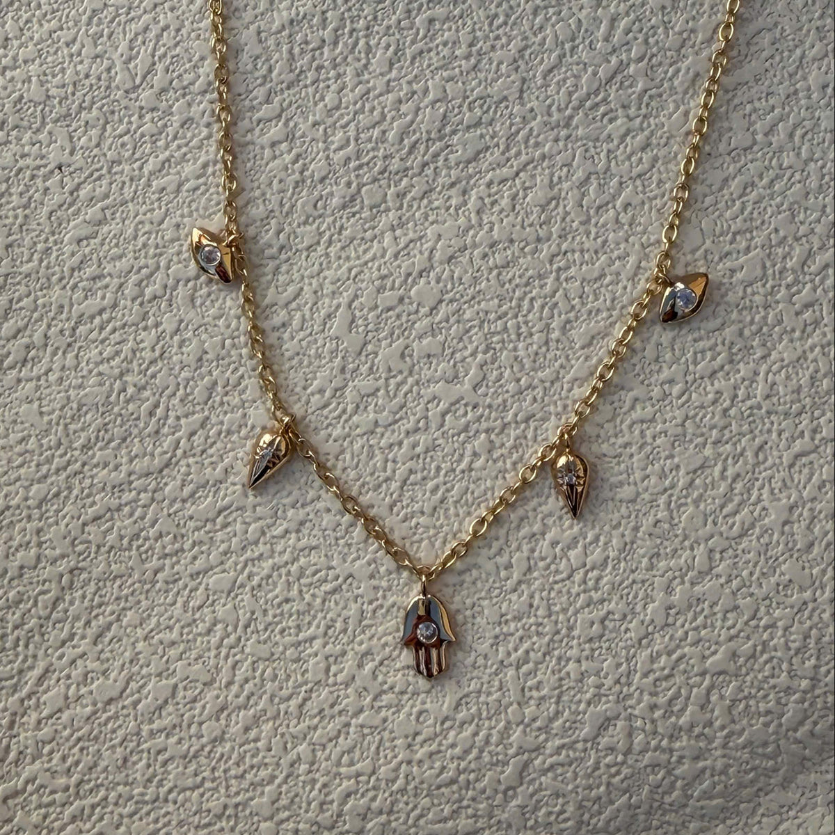 A delicate gold chain necklace featuring small dangling charms, including a central hamsa hand pendant with a blue stone, laid flat on a textured light gray fabric background.