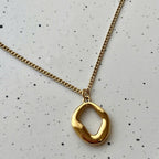 A gold chain necklace with a polished, abstract open oval pendant, displayed on a light speckled white surface.