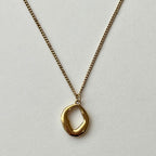 A gold chain necklace with a polished, abstract oval pendant, displayed against a clean, light gray textured background.