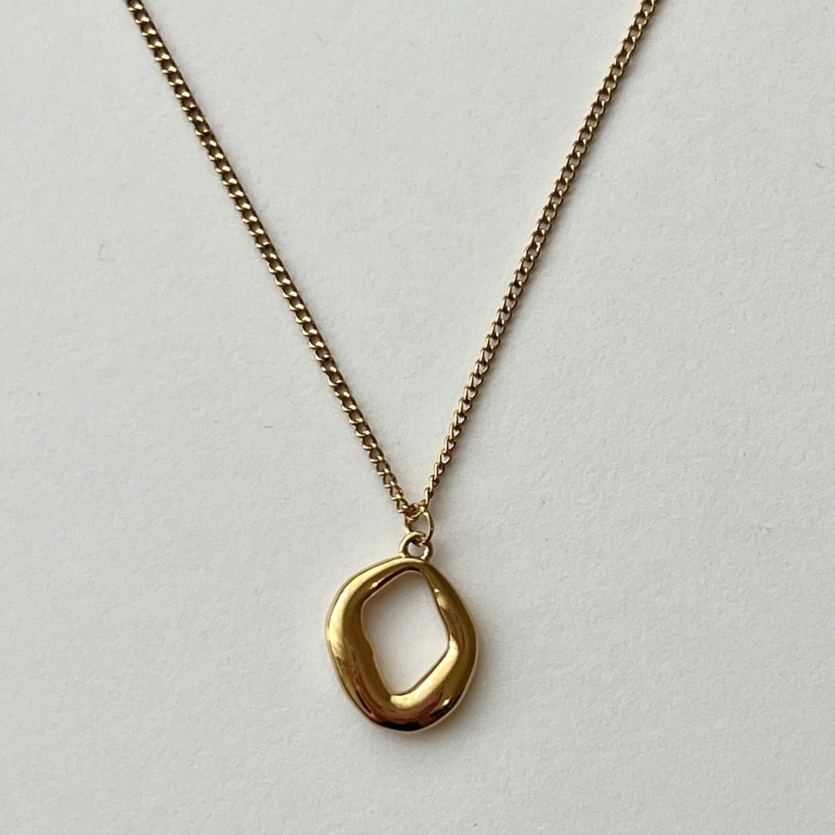 A gold chain necklace with a polished, abstract oval pendant, displayed against a clean, light gray textured background.