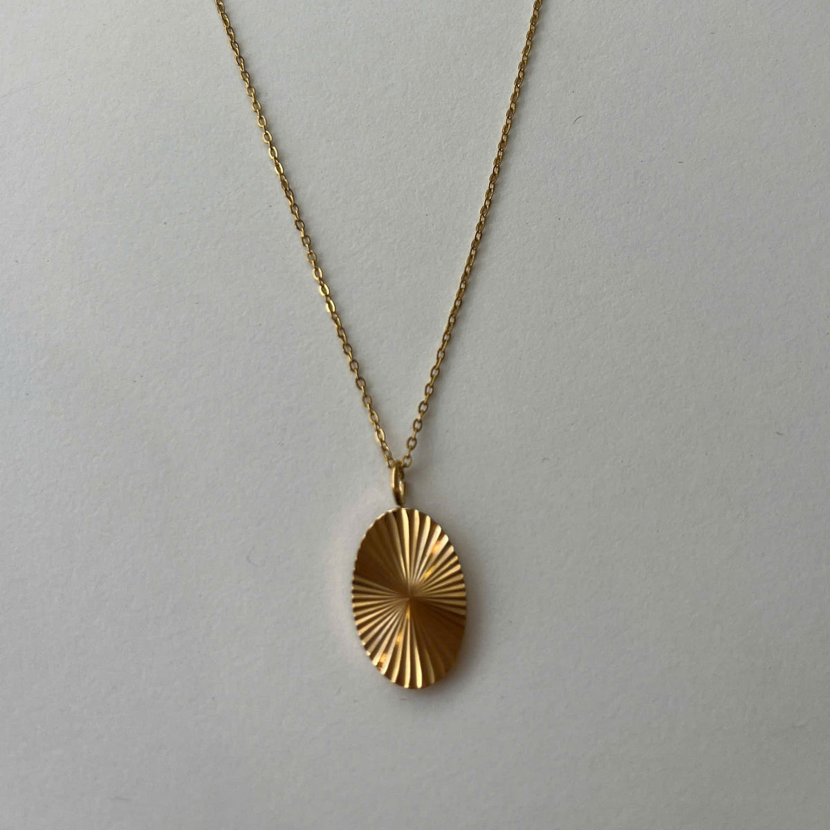 A gold chain necklace with a textured, sunburst-style oval pendant, displayed against a plain light gray background.