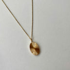 A gold chain necklace with a textured, sunburst-patterned oval pendant, displayed on a neutral light gray background.