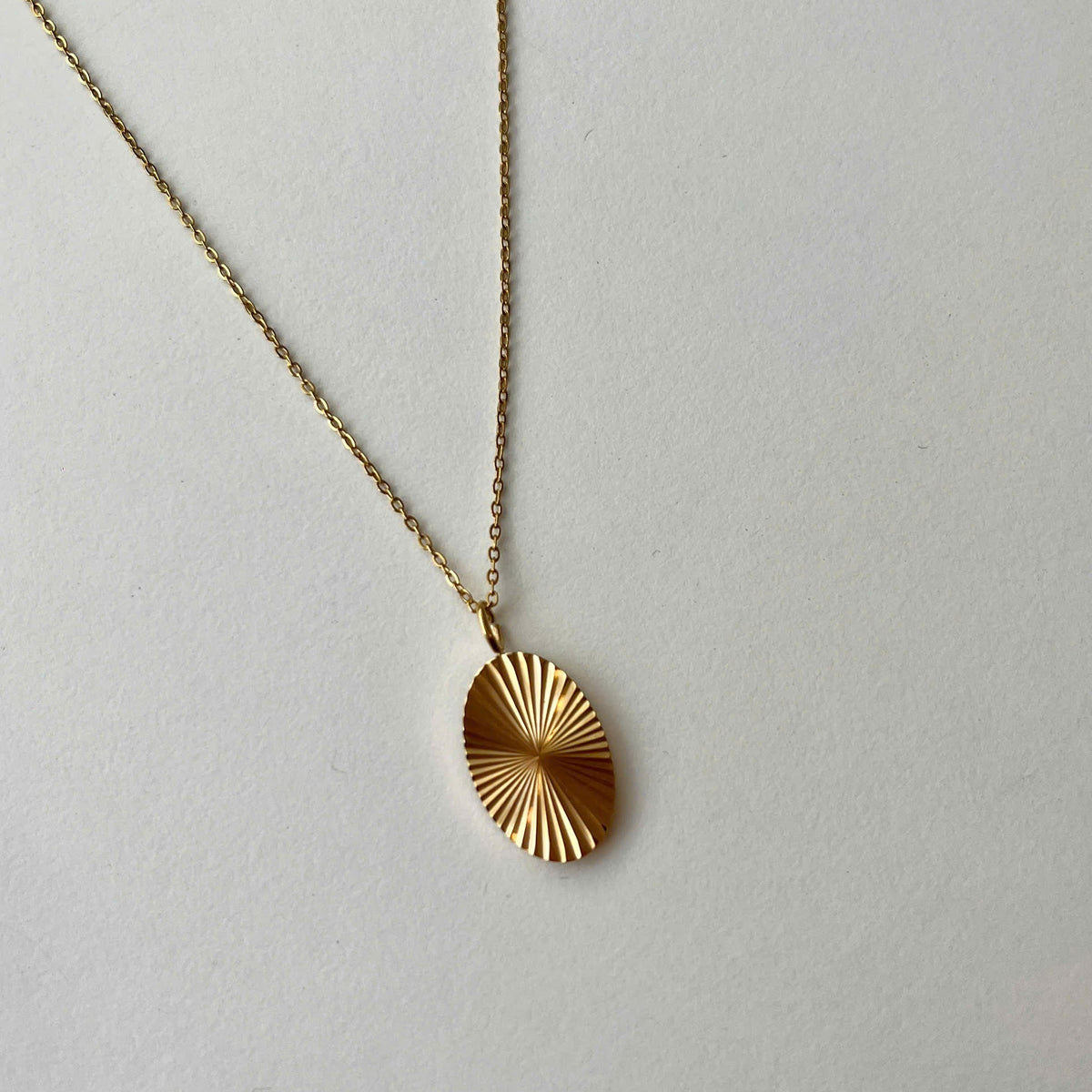A gold chain necklace with a textured, sunburst-patterned oval pendant, displayed on a neutral light gray background.