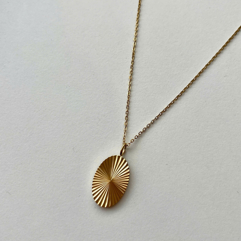 A gold oval pendant necklace with a sunburst or radial textured pattern, hanging from a delicate gold chain against a plain off-white background.