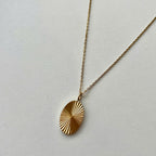 A gold oval pendant necklace with a sunburst or radial textured pattern, hanging from a delicate gold chain against a plain off-white background.