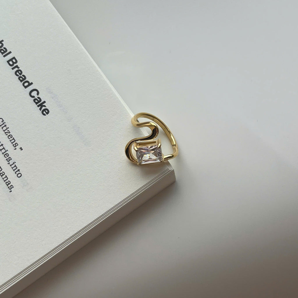 A gold-plated ring with a clear rectangular gemstone, resting on the edge of a white book against a light gray background.