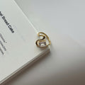 A gold-plated ring with a clear rectangular gemstone, resting on the edge of a white book against a light gray background.