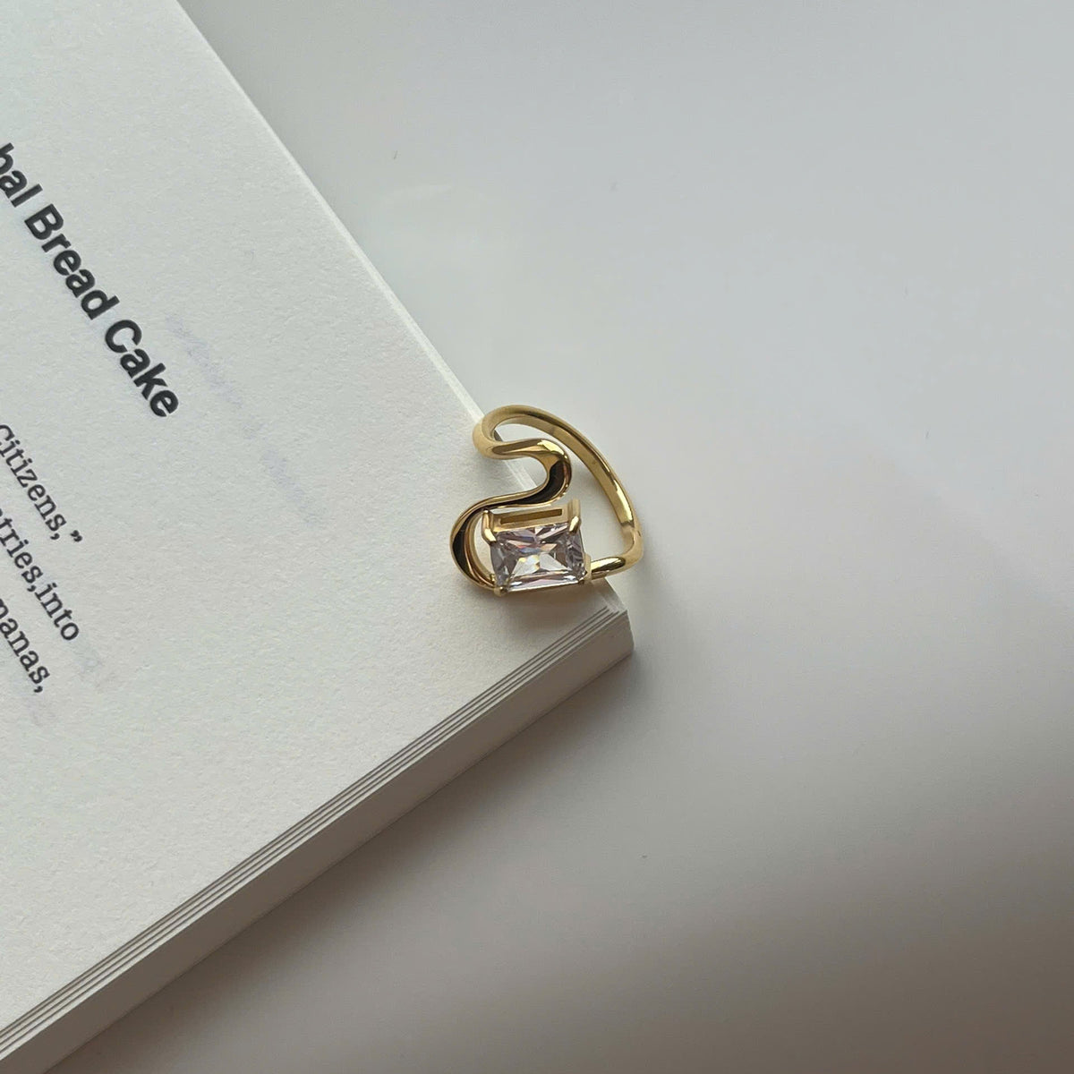 A gold-plated ring with a clear rectangular gemstone, resting on the edge of a white book against a light gray background.