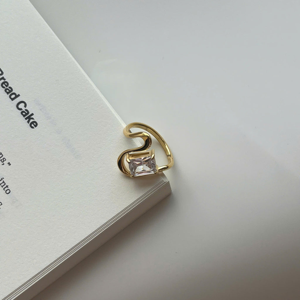 A gold-plated ring with a rectangular clear gemstone, resting on the edge of a white book against a light gray background.