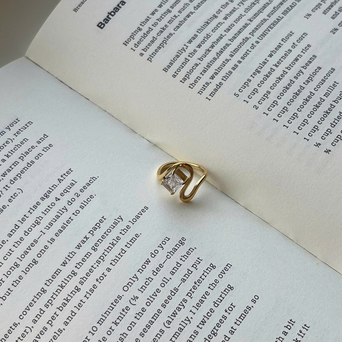 A gold ring with a clear rectangular gemstone resting on an open book with printed text.