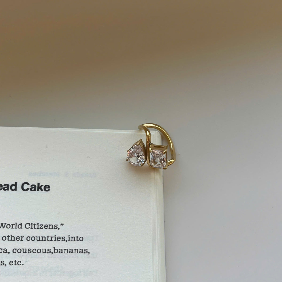A gold crescent-shaped ring with two clear triangular gemstones, resting on the edge of an open book page.