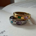 Two rings stacked on an open book: one gold ring with a green rectangular gemstone and pink marquise gemstone, and one silver ring with a purple marquise gemstone, a blue round gemstone, and small clear stones.