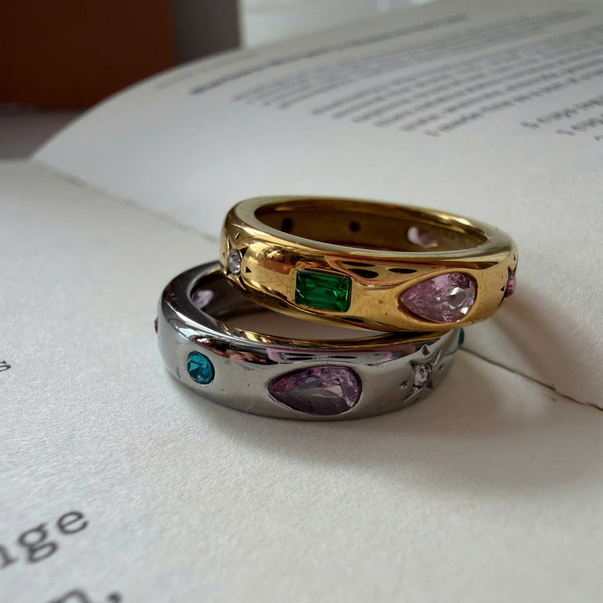 Two rings stacked on an open book: one gold ring with a green rectangular gemstone and pink marquise gemstone, and one silver ring with a purple marquise gemstone, a blue round gemstone, and small clear stones.