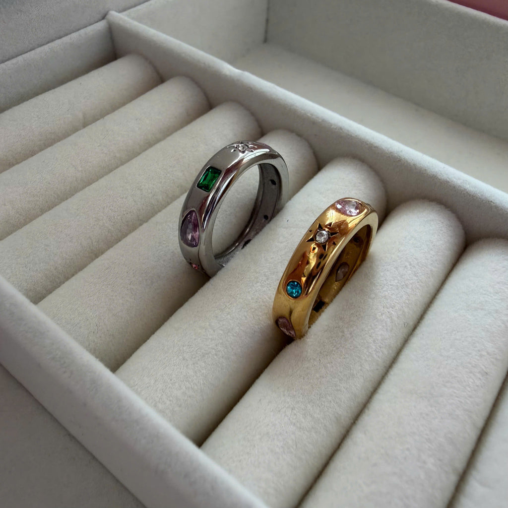 Two rings displayed in a cream-colored jewelry box: one silver band with green and purple stones, and one gold band with pink, blue, and green stones.