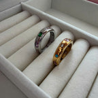 Two rings displayed in a cream-colored jewelry box: one silver band with green and purple stones, and one gold band with pink, blue, and green stones.