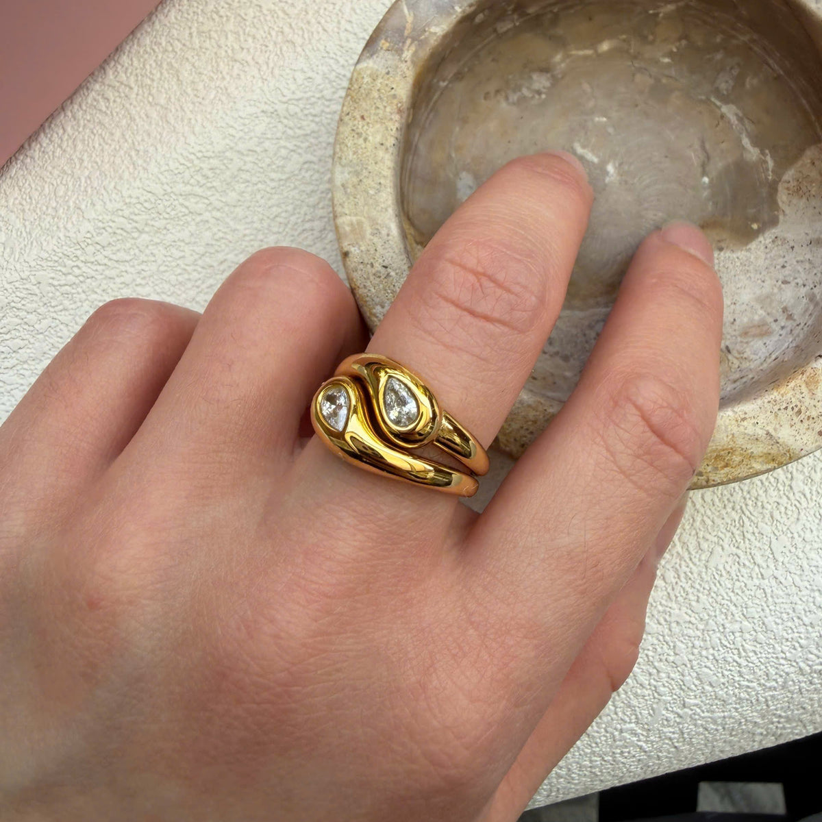 A gold ring with two heart-shaped clear gemstones worn on a person's finger, shown in a close-up shot next to a stone dish.