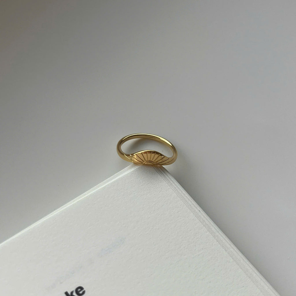 A delicate gold ring with a small sunburst or fan-like motif on the front, resting on the corner of a white book against a light gray background.