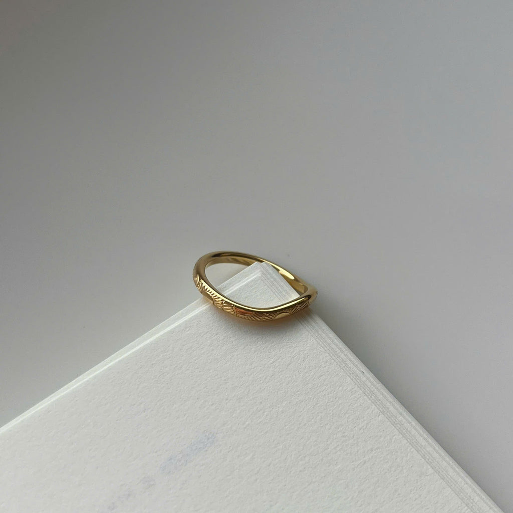 A thin gold ring with a textured, slightly wavy band, resting on the corner of a white textured card against a neutral gray background.