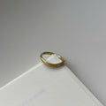 A thin gold ring with a textured, slightly wavy band, resting on the corner of a white textured card against a neutral gray background.