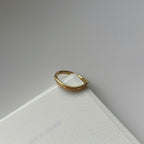 A thin gold ring with a textured, slightly wavy band, resting on the corner of a white textured card against a neutral gray background.