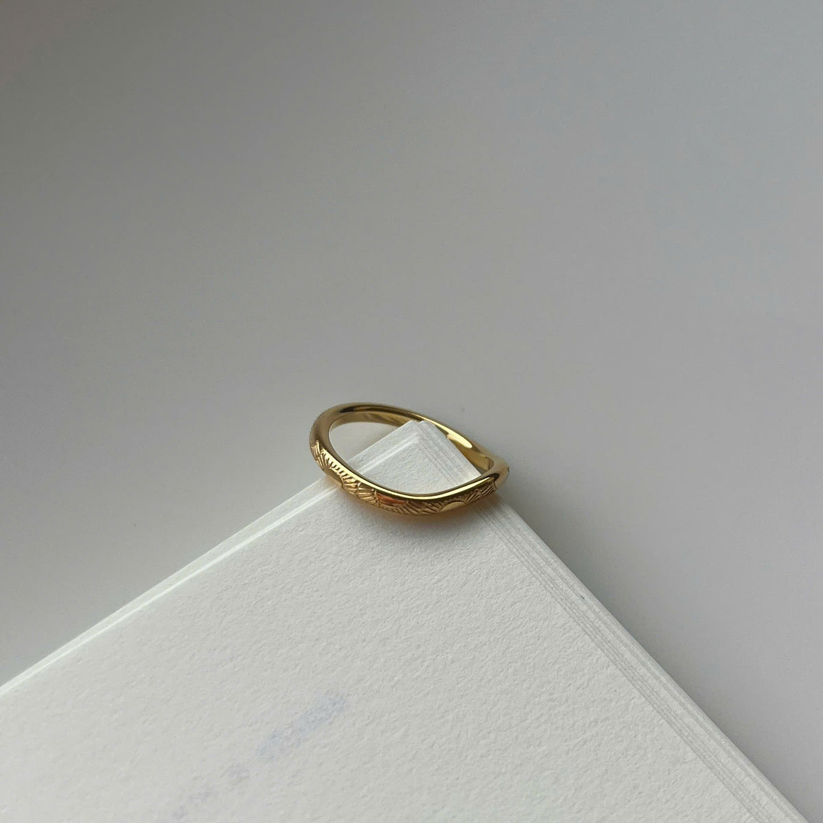 A thin gold ring with a textured, slightly wavy band, resting on the corner of a white textured card against a neutral gray background.