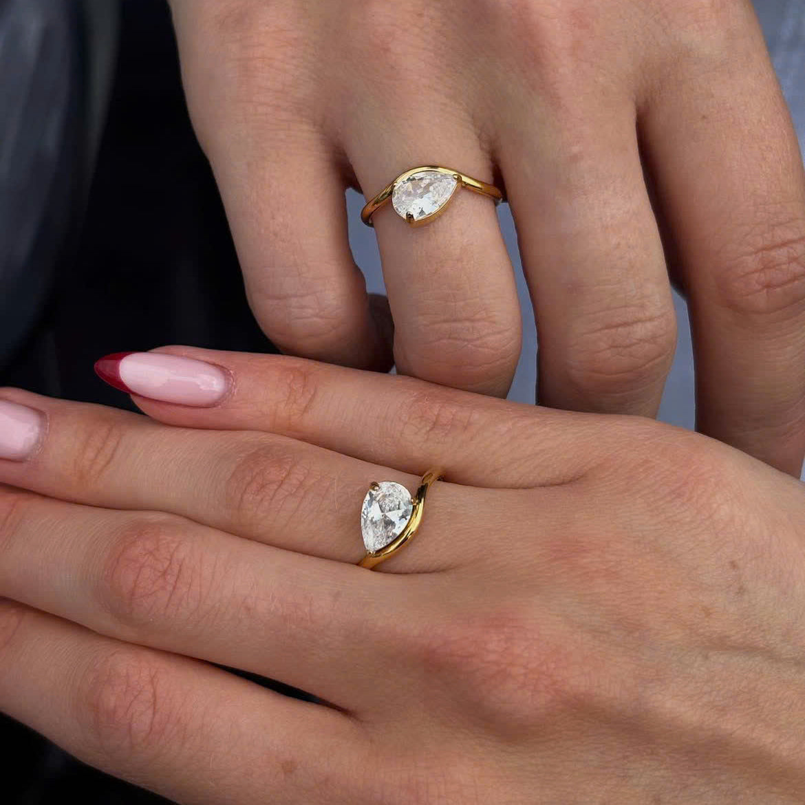 Two hands shown wearing gold rings with pear-cut clear gemstones, one larger ring on a higher finger and one smaller ring on a lower finger.
