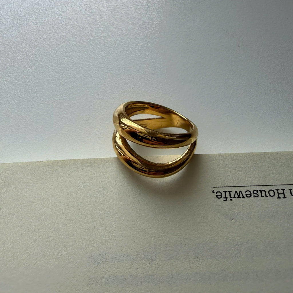 A gold-toned twisted ring with three interlocking bands, resting on the edge of an open book against a light gray background.