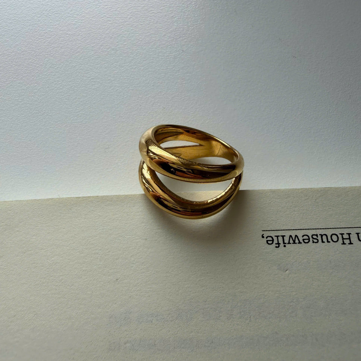 A gold-toned twisted ring with three interlocking bands, resting on the edge of an open book against a light gray background.