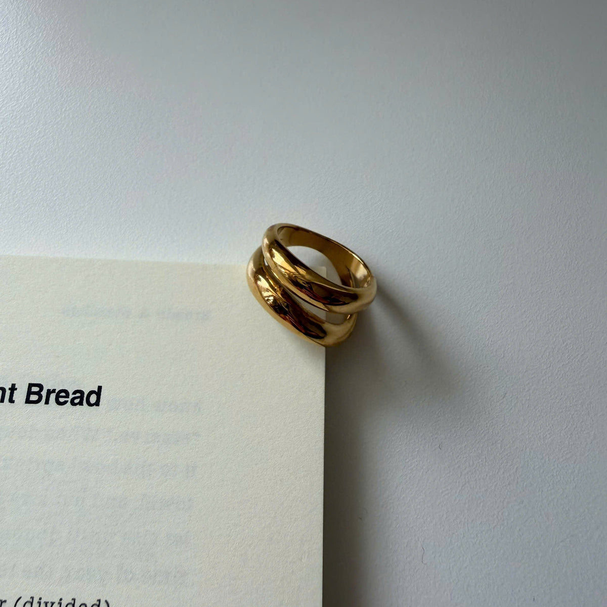 A gold double-band ring with a polished, reflective finish resting on the corner of an off-white book page against a neutral gray background.