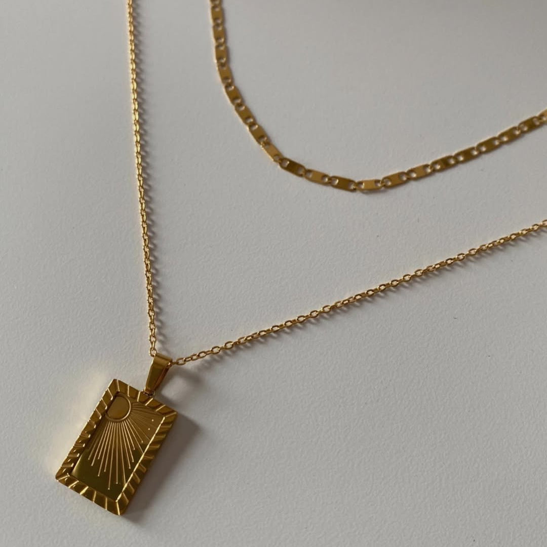 A gold-toned necklace with a rectangular pendant featuring a sunburst design, displayed on a light gray background with a second chain draped behind it.